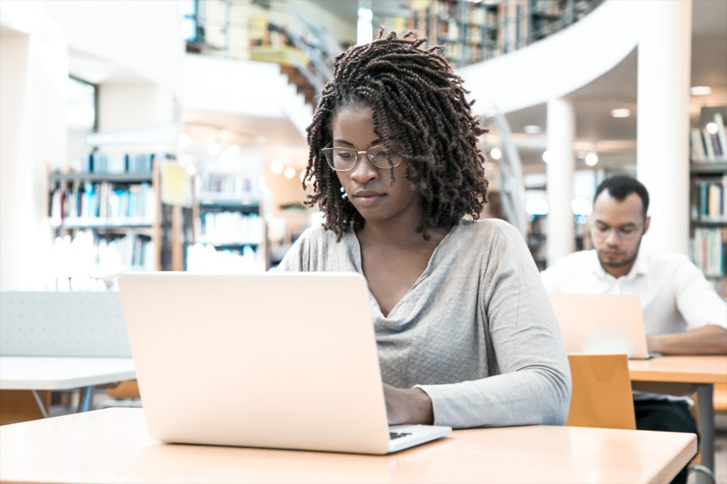 student working on research paper in library. People sitting at desks and using laptops in computer class with bookshelves.
